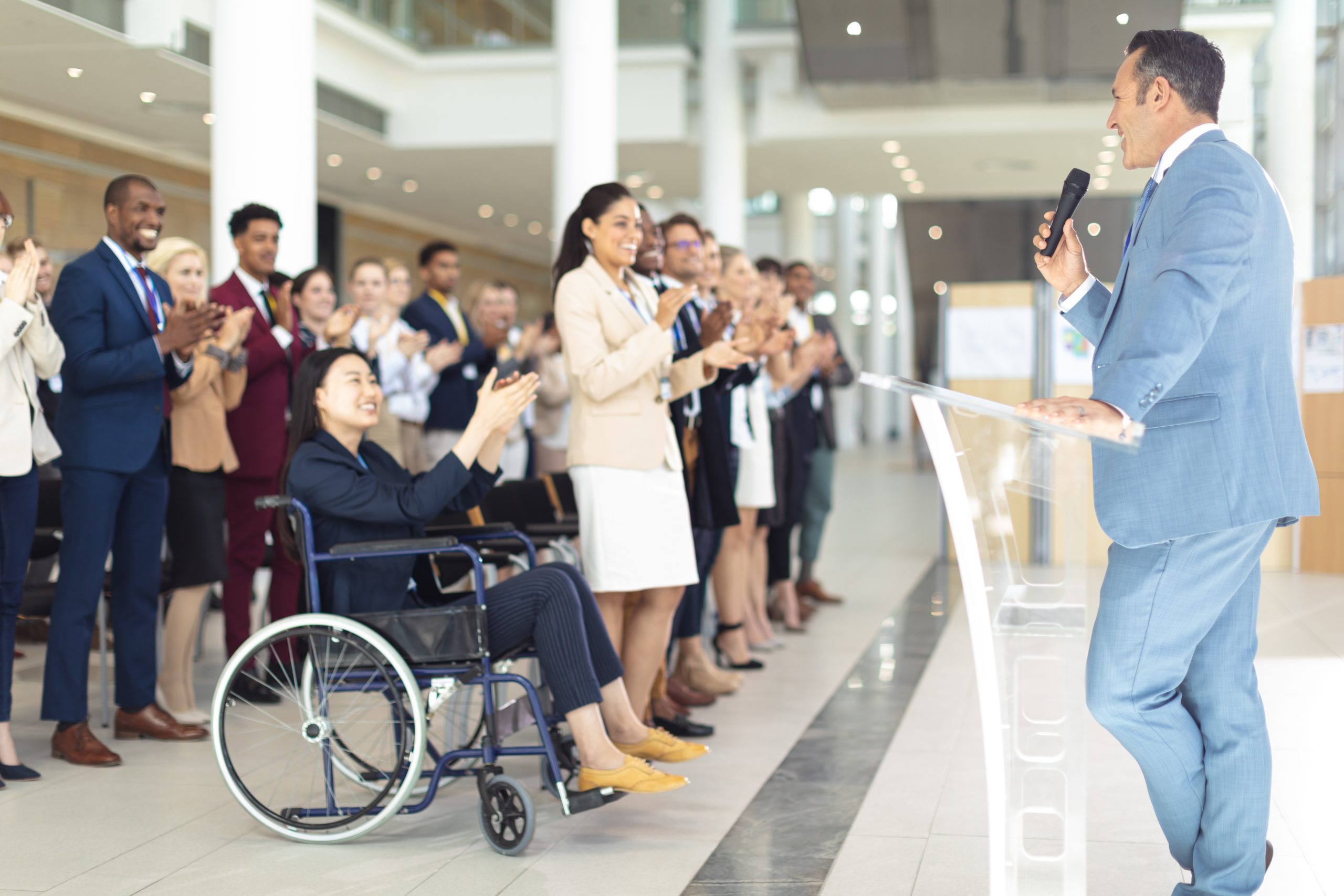 A diverse group of professionals, including a woman in a wheelchair, applauding a keynote speaker at an inclusive business conference in a modern venue.